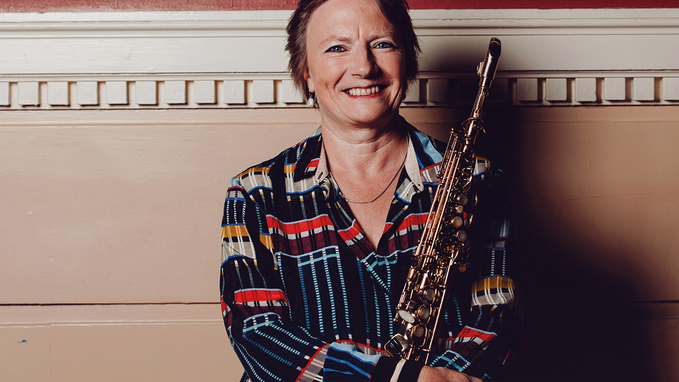Jo stands against a wall holding a saxophone and smiling
