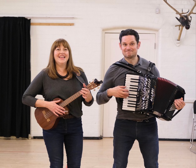 Emmie playing a banjo and Nicholas playing an accordion, both looking into the camera and smiling 