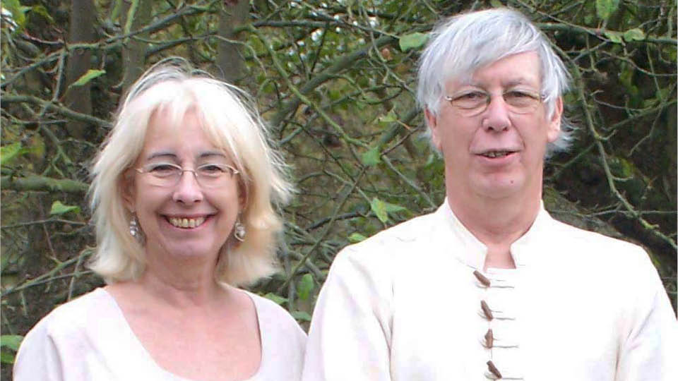 couple wearing white tops, standing side by side facing the camera, with trees close behind