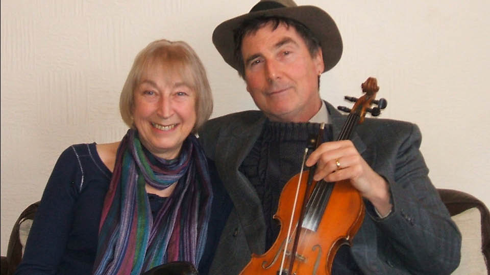smiling seated couple looking at camera, he is holding a fiddle