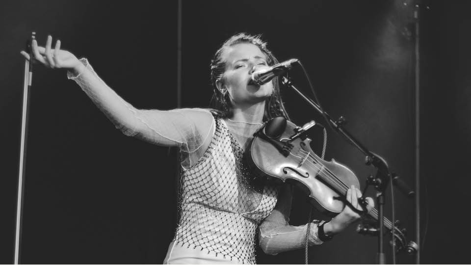 Woman on stage in white sleeveless top, singing into microphone and holding fiddle
