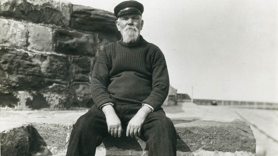 A black-and-white photo of a bearded man on a harbour wall.