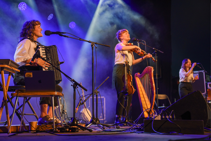A trio of singers on stage, singing into microphones. One is also playing accordion, and another is also playing the fiddle.