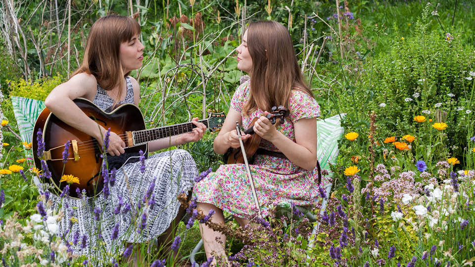 Two women with fair hair (the Carrivick sisters) sit amongst wild flowers, singing to each other and holding a guitar and a fiddle